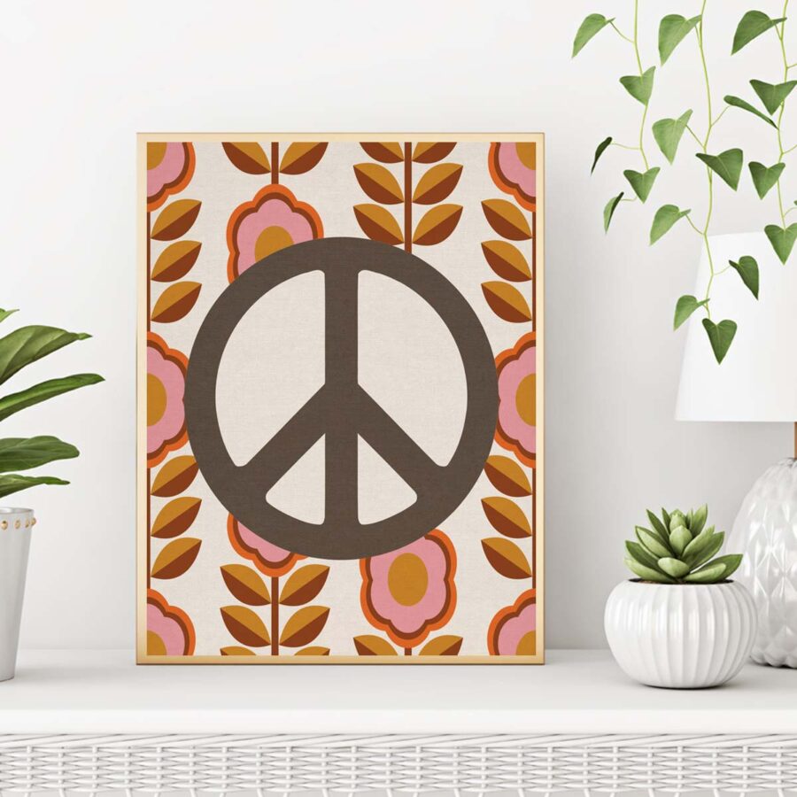01-1-peace-symbol-display-3 A framed artwork with a large dark brown peace symbol centered on a cream background. Surrounding the peace sign are symmetrical retro-style floral patterns, featuring pink, orange, and brown flowers and leaves. The frame rests on a white surface, with a potted green succulent plant to the right and a white lamp with a glass base in the background. On the left side, part of another green plant is visible.