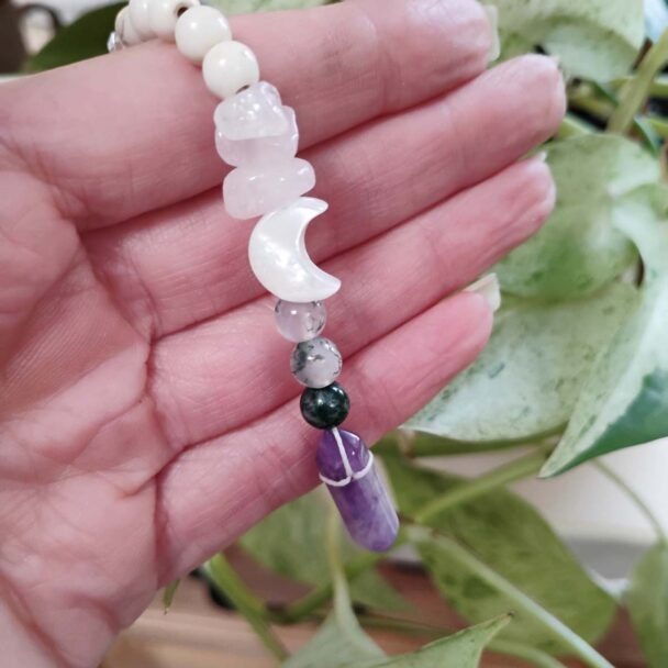 A hand holding a gemstone charm with rose quartz chips, a white crescent moon bead, several round stone beads, and a purple amethyst crystal point, with green leaves in the background.