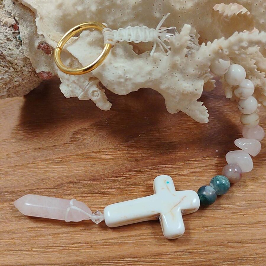 rose-quartz-crystal-faith -car -charm -liteorb Close-up of a handmade car charm showing a white stone cross, a rose quartz crystal point, several small stone beads, and a gold metal ring, resting on a wooden surface.
