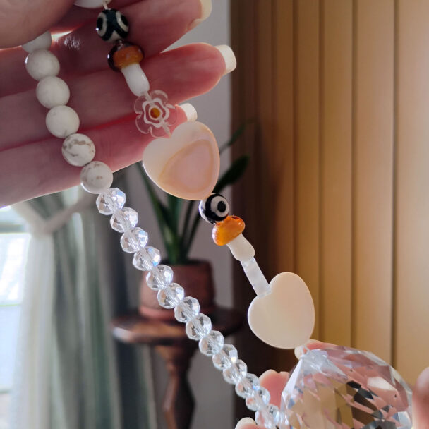 Close-up of a handmade car charm showing a white stone cross, a rose quartz crystal point, several small stone beads, and a gold metal ring, resting on a wooden surface.