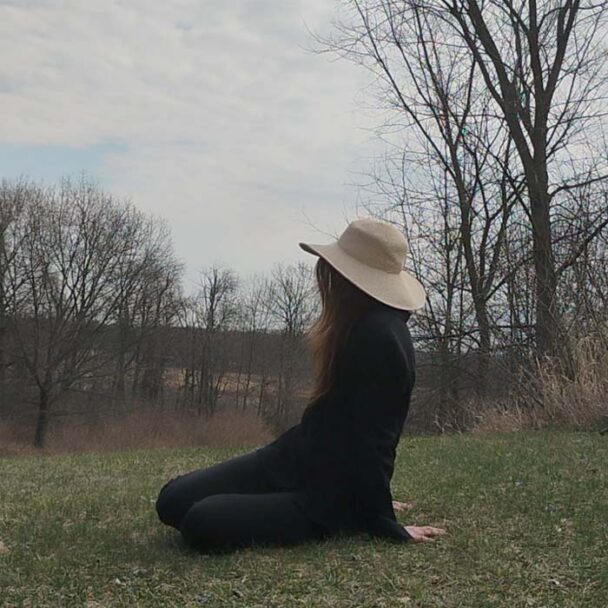 woman sitting on the ground dressed in black , wearing a straw hat looking over a field.